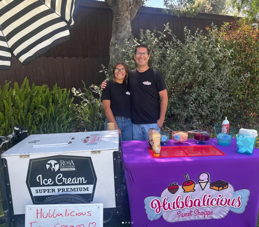 Two people standing behind a purple ice cream stand with 'Hubbalicious Sweet Shoppe' branding.