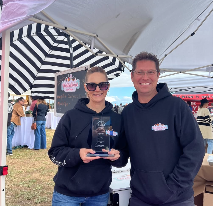 Two people in navy blue hoodies with a logo, one holding an award, standing under a tent with striped umbrella.